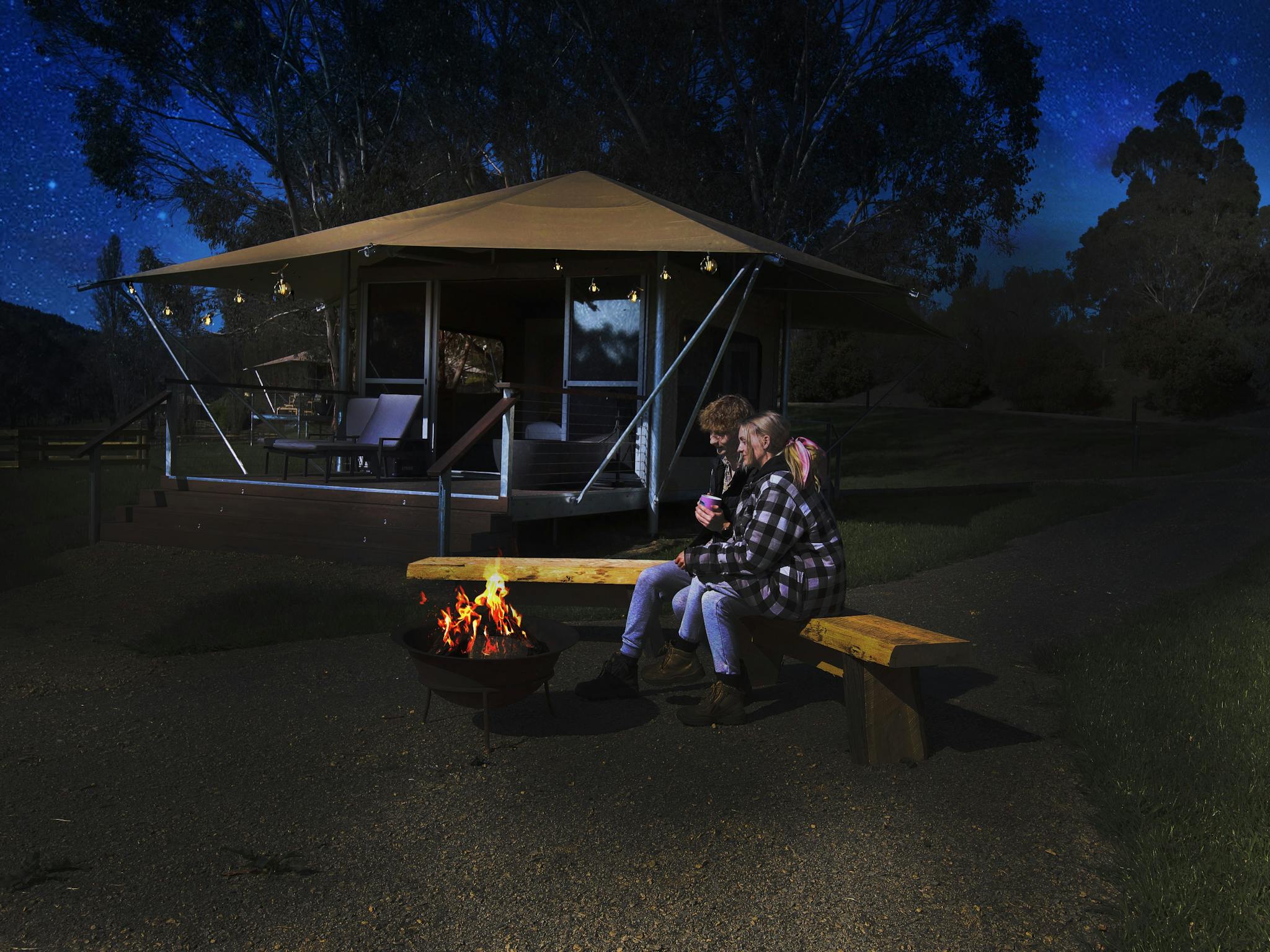 A couple sitting around a fire pit at night with eco glamping tent in the background