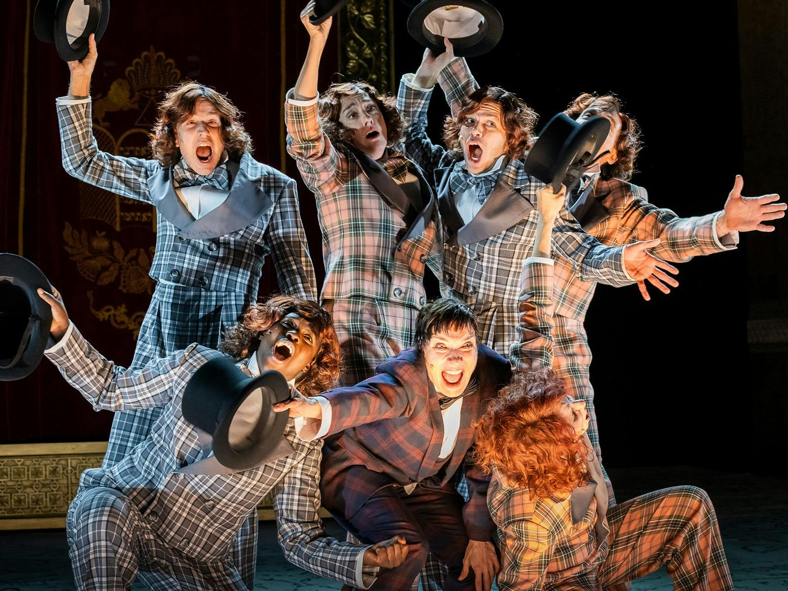 Actors on stage wearing plaid suits pose with their arms out holding top hats