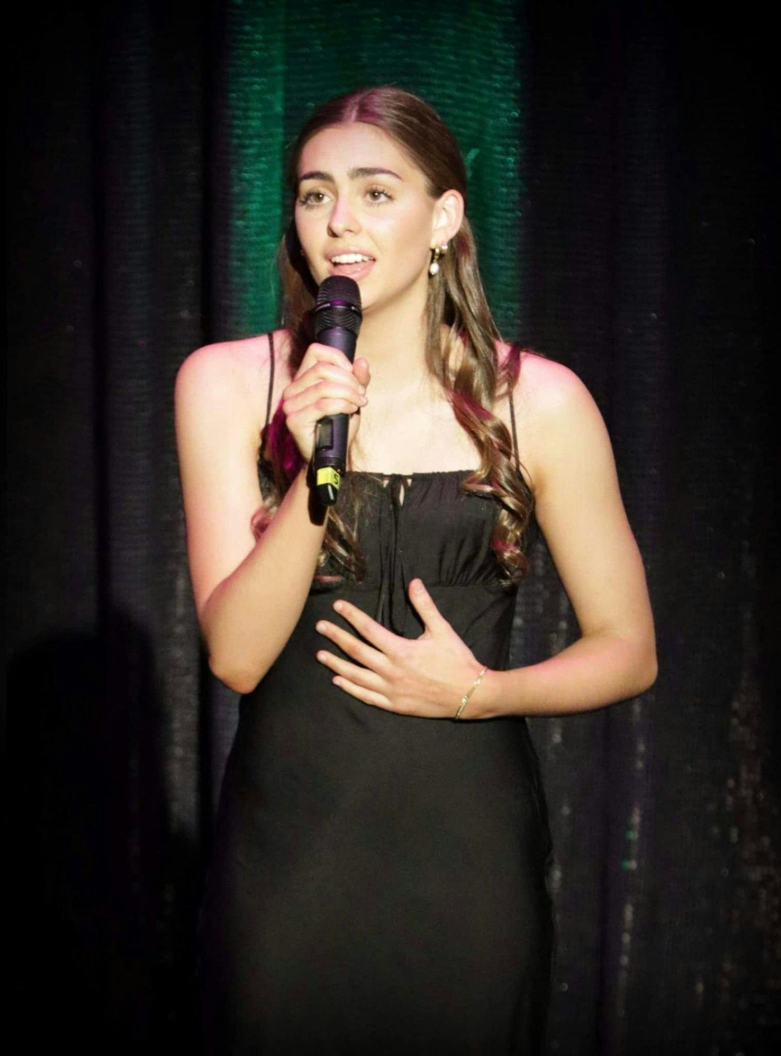 A lady singing into a microphone, left arm on her stomach, wearing a black dress.