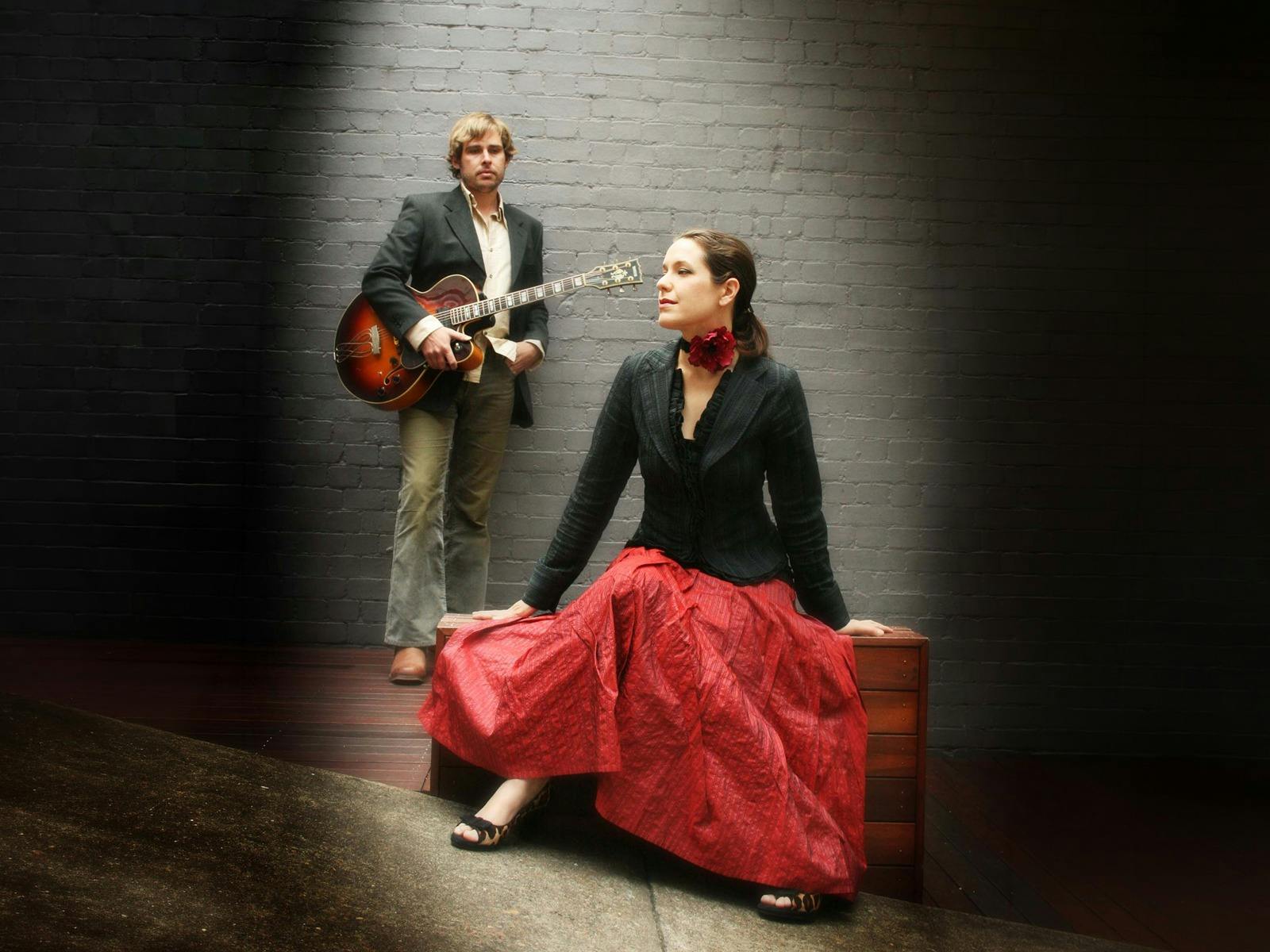 A man holds a guitar and leans against a wall, a woman in a red skirt sits on a timber box