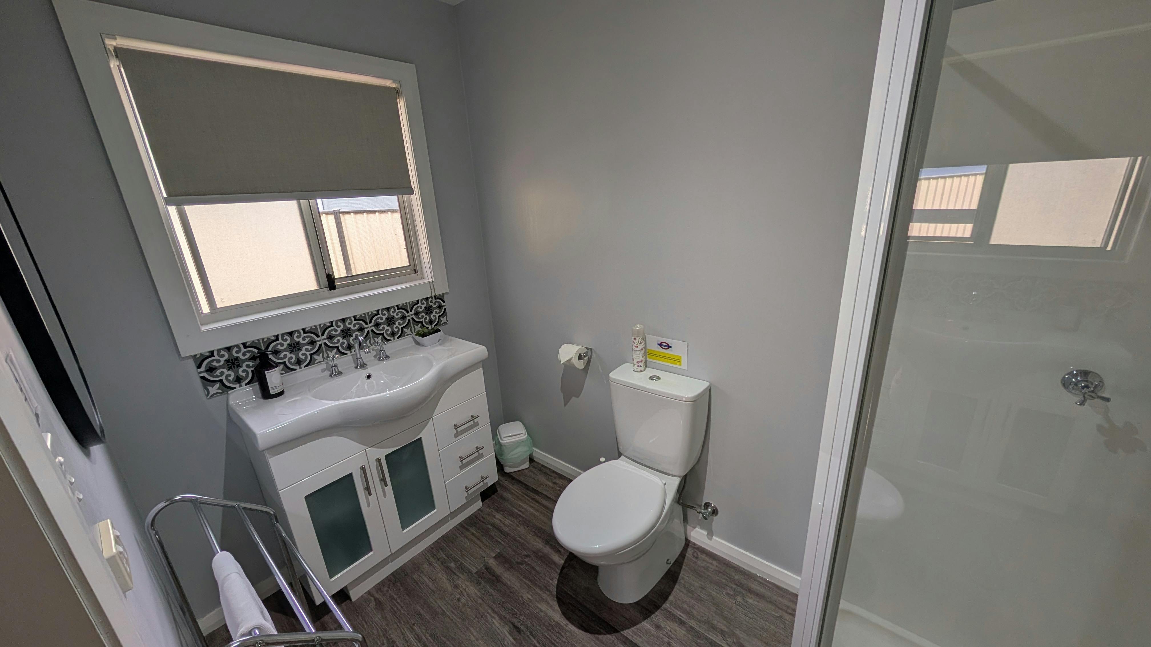 Bathroom with vanity, toilet, shower, towel rack and window with blind.
