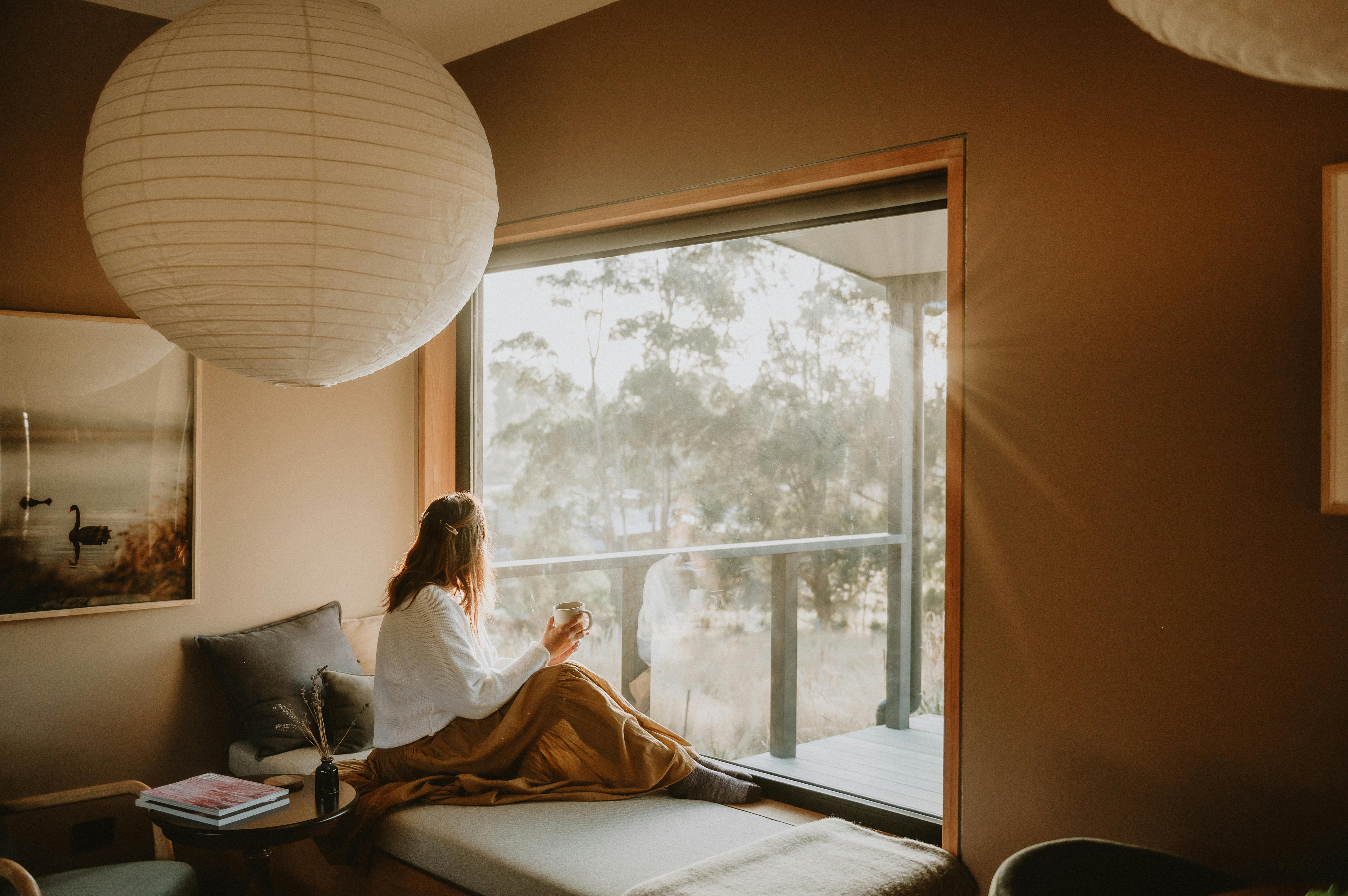A lady holding a mug and looking out a large window