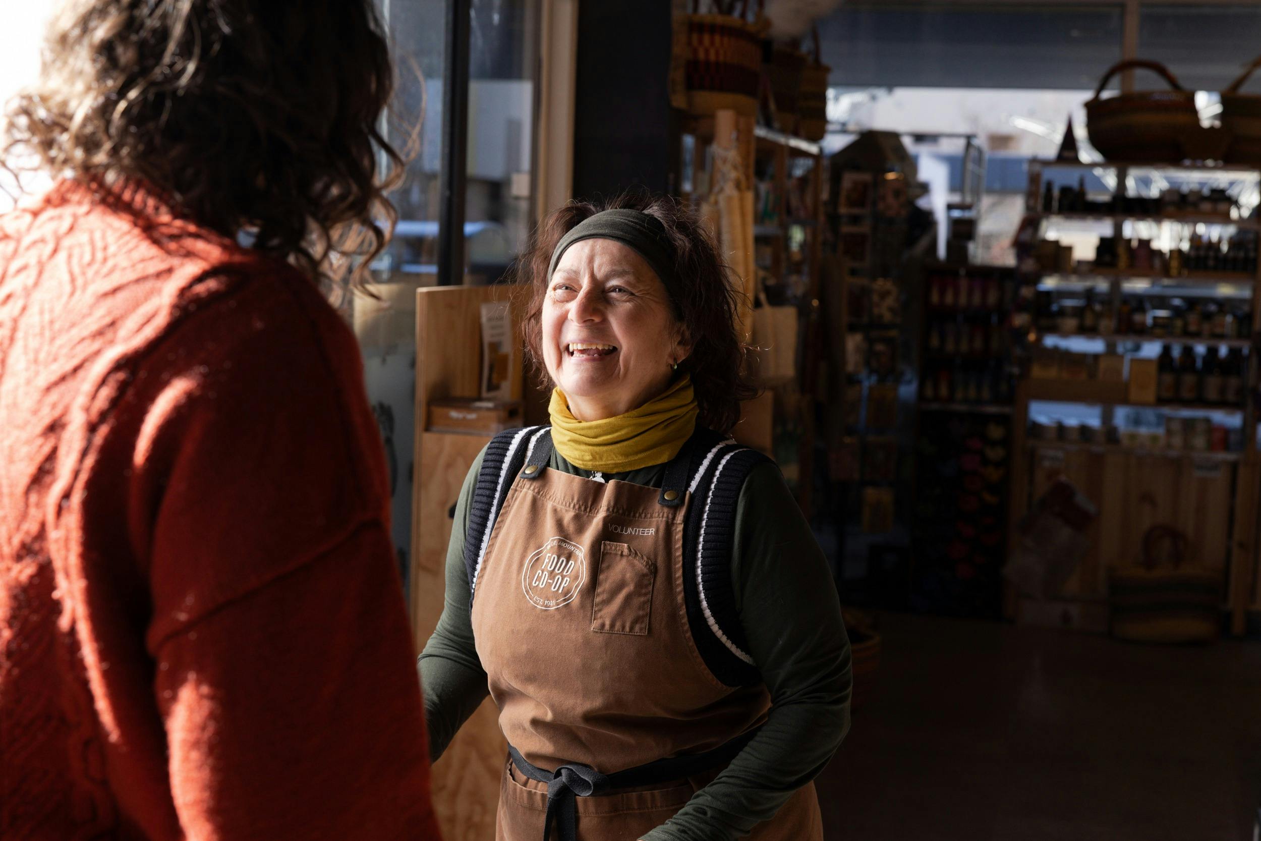 Smiling woman in a brown volunteer apron faces another person, back towards camera.