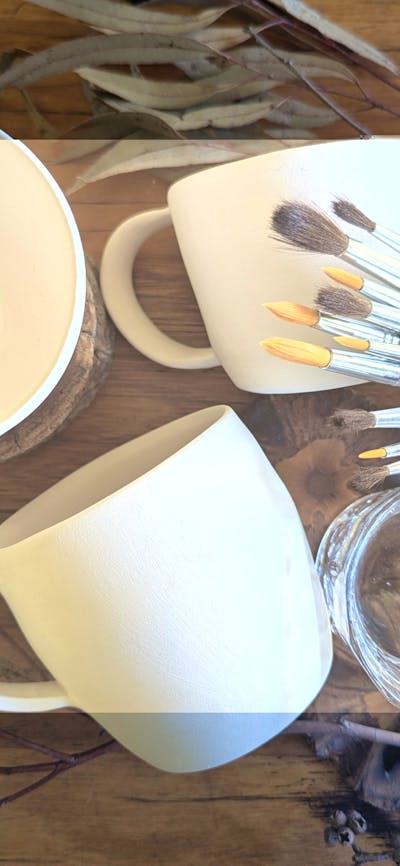 Ceramic mugs awaiting glazing with paint brushes and gin and tonic