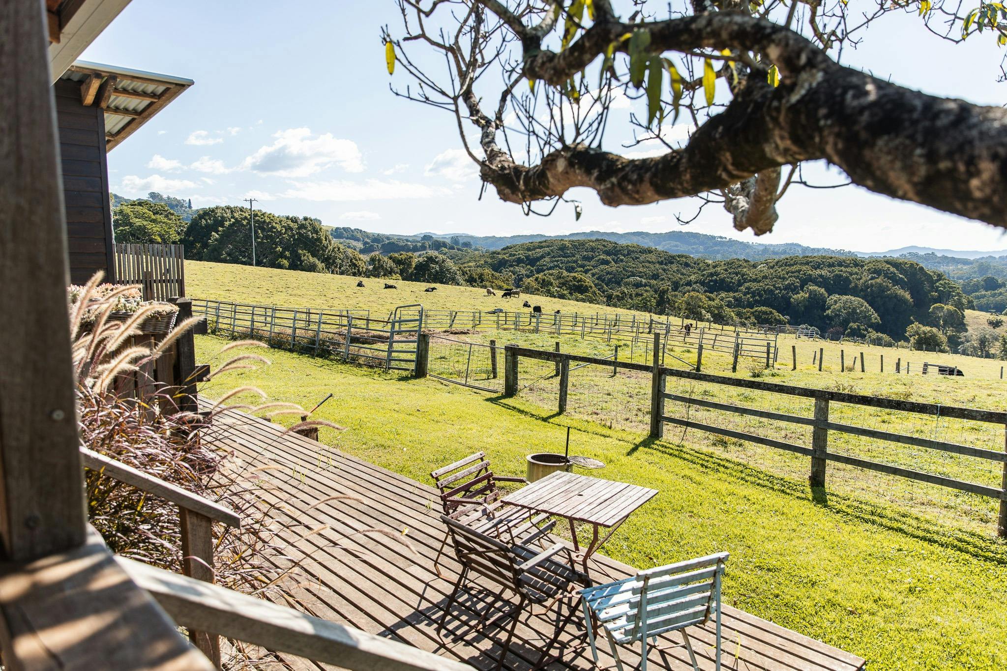 Two chairs and a small table sit on a timber deck overlooking a farm and valley on a sunny day