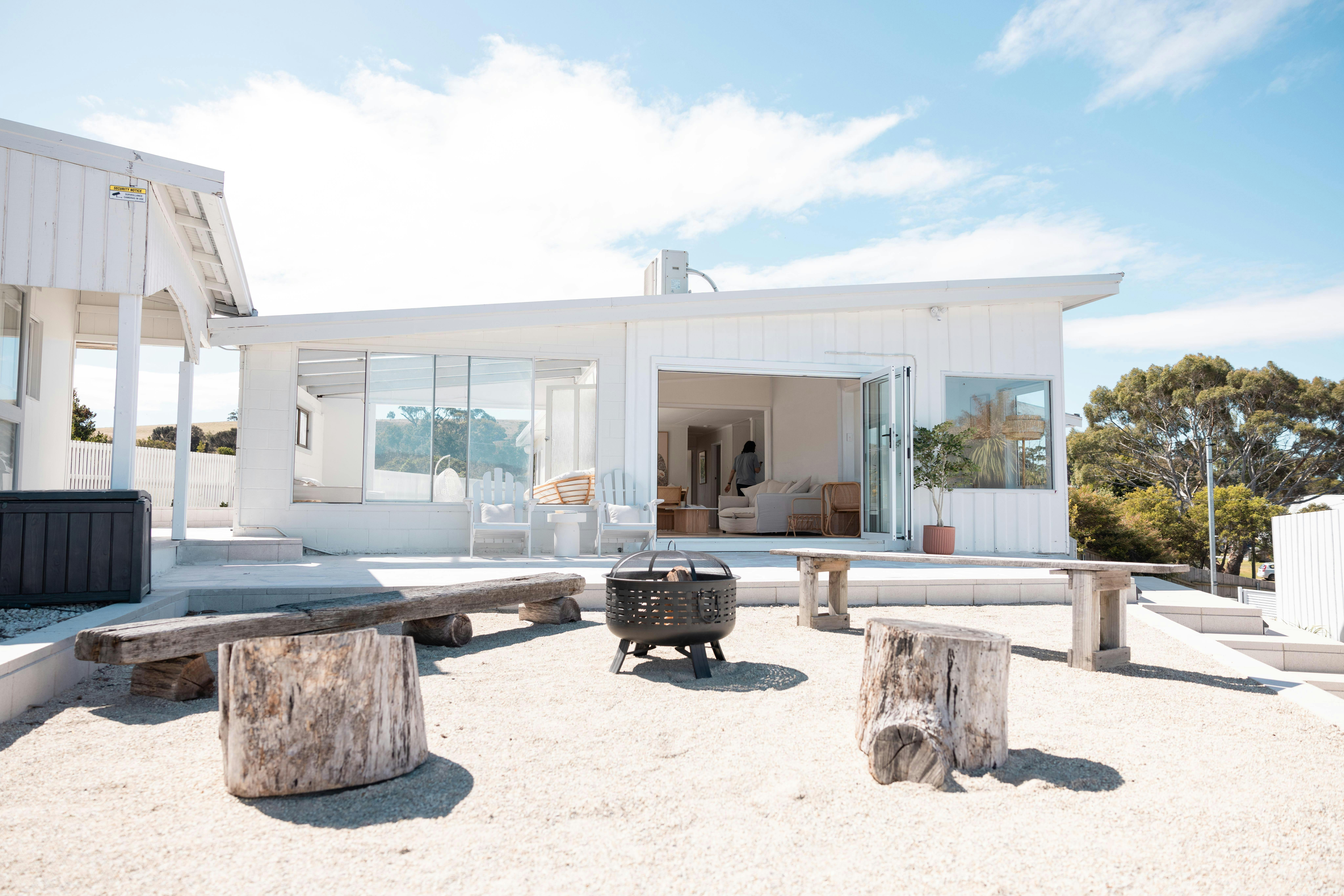 Exterior view of a white beach house with an open patio area, fire pit, and rustic wooden seating