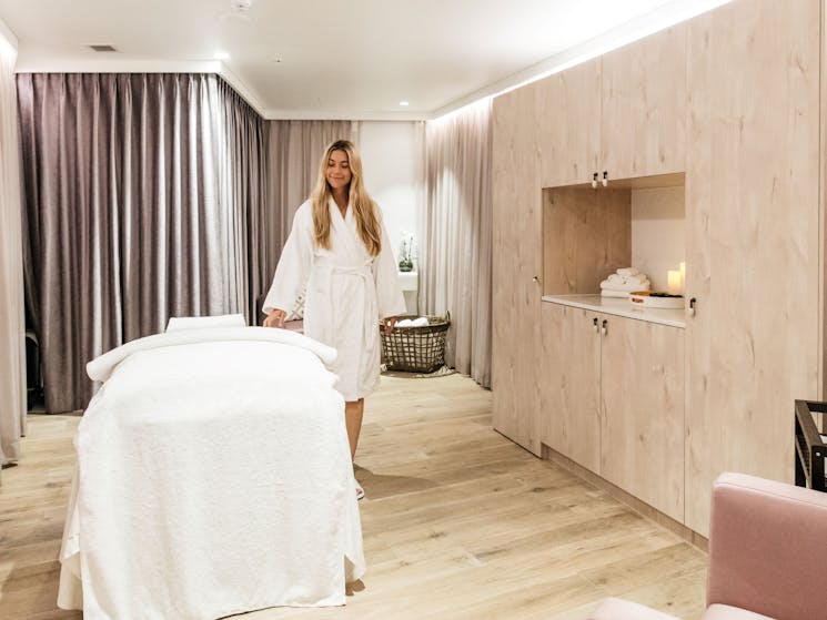 A woman stands next to a massage table dressed in a robe inside a treatment room