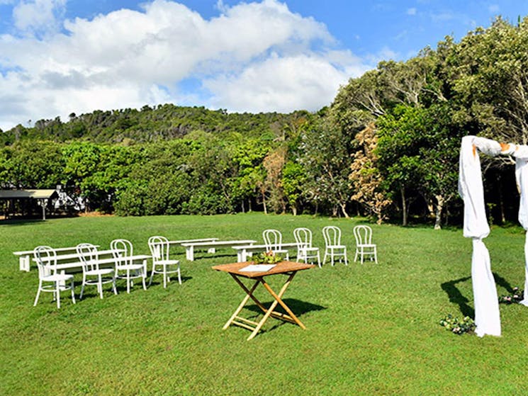 Chairs, a table and a bridal arch set up on Broken Head lawn with lush rainforest in the background