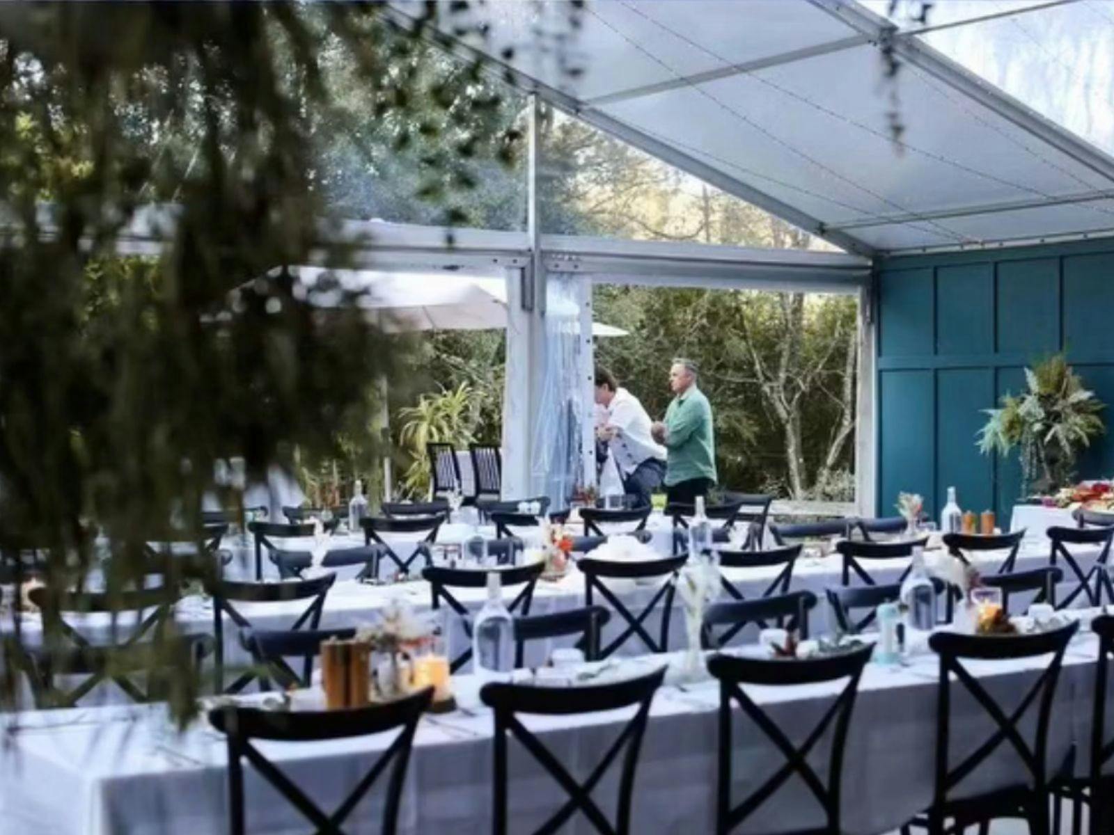 Outdoor marquee with long tables, black chairs, white tablecloths, and greenery in the background.