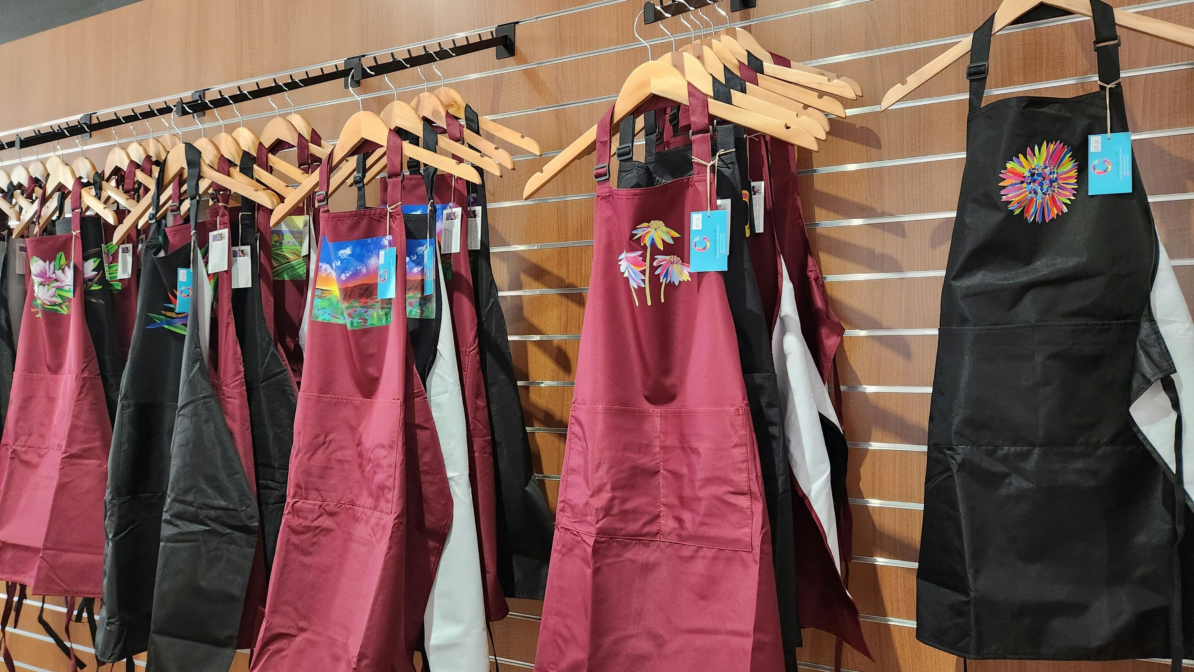 Red and black aprons  with colourful designs hung on rows of clothe racks.