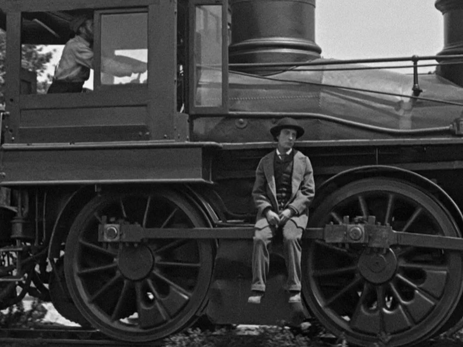 Buster Keaton sits looking dejected on the side rod of a steam locomotive as it slowly drives away.