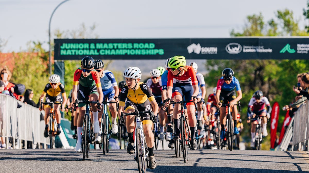 A peloton of junior road cyclists pass under the finish arch during a National Championship race.