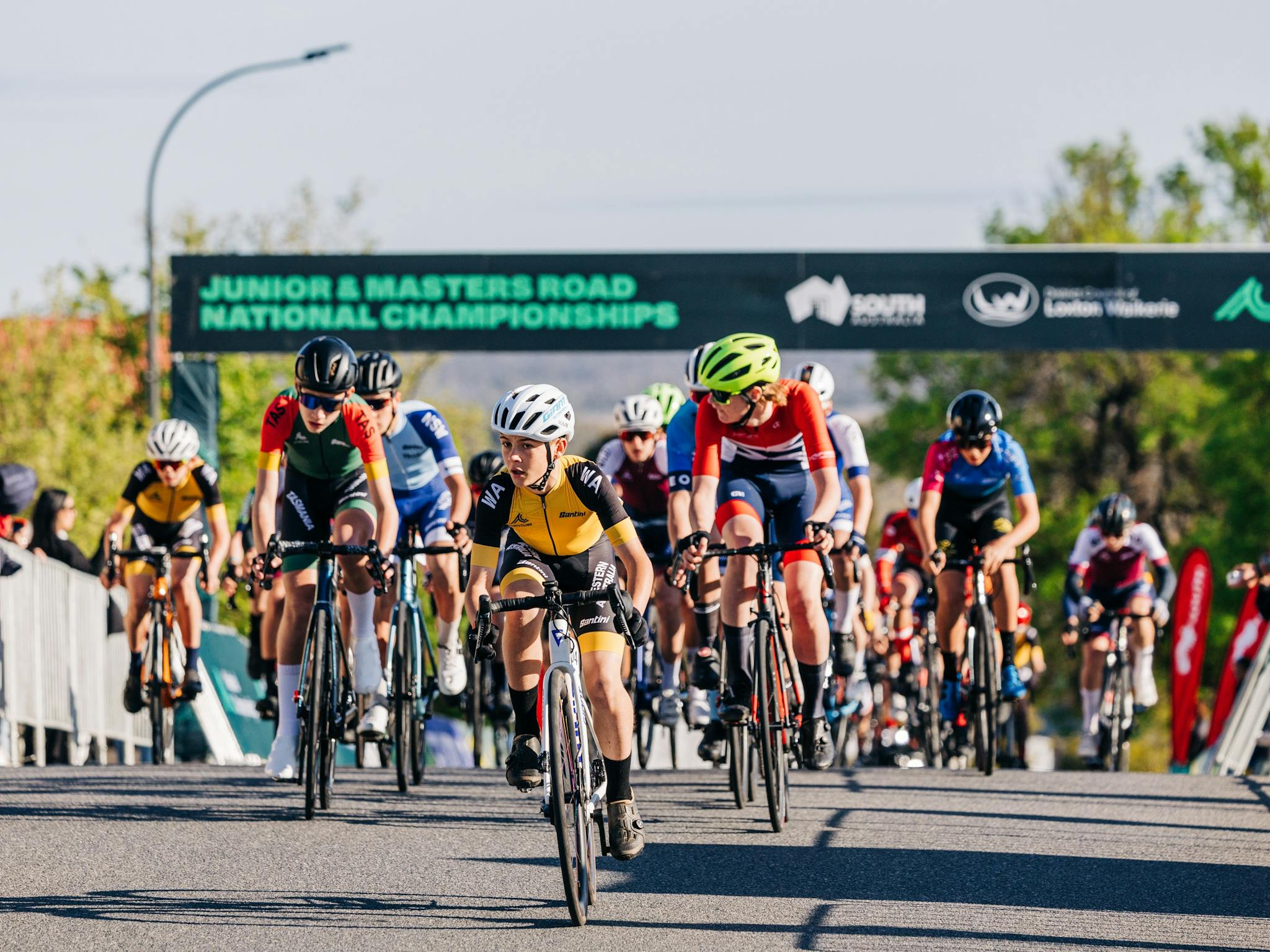 A peloton of junior road cyclists pass under the finish arch during a National Championship race. A peloton of junior road cyclists pass under the finish arch during a National Championship race.