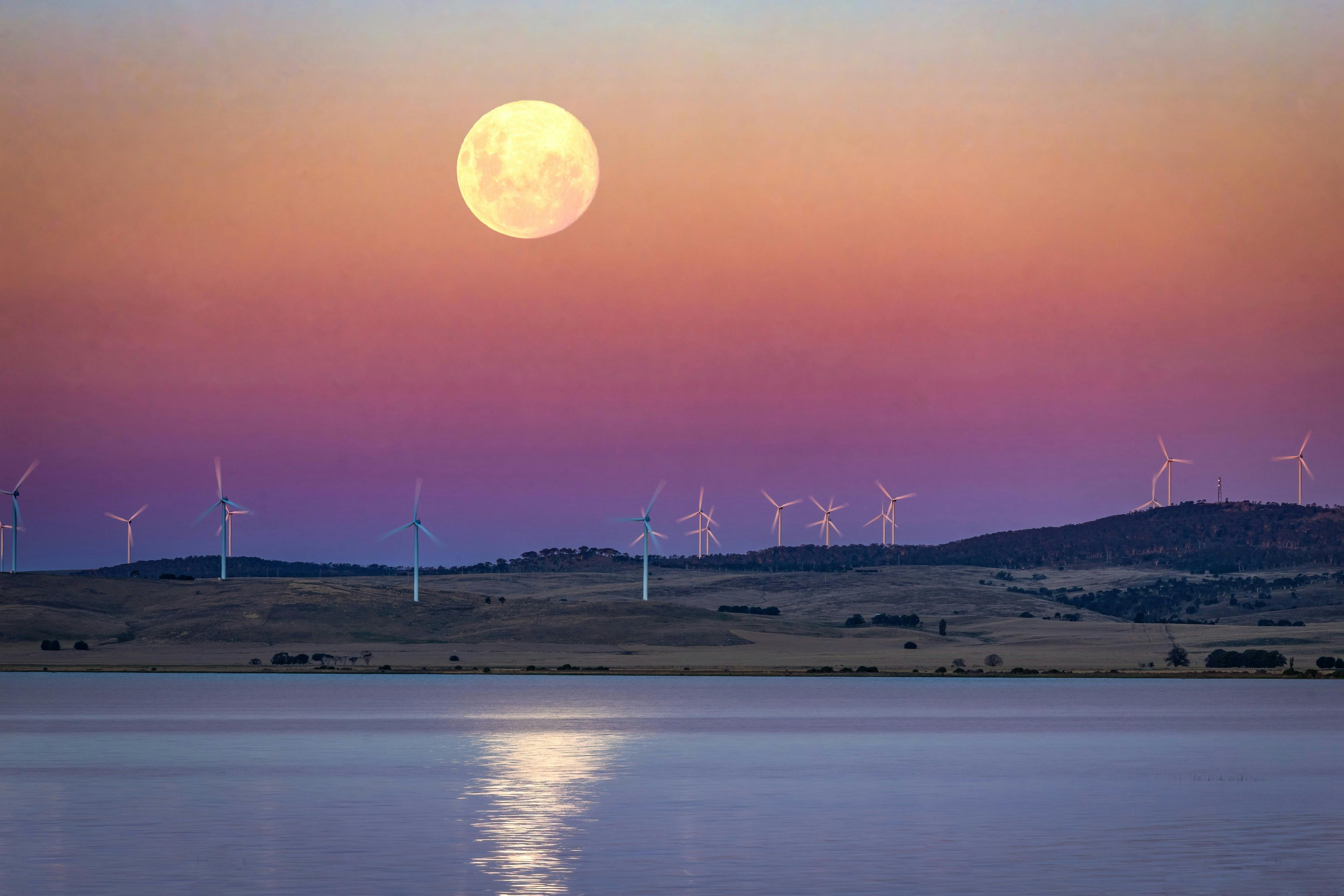 A large golden full moon rises over a calm lake at dusk, beneath a pink and purple sky with a distan