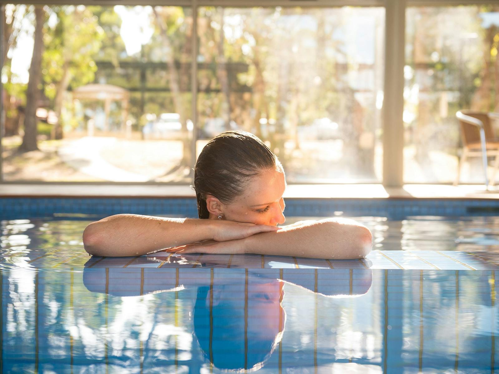 Young woman in pool resting head on her hands on the ledge to the spa with daylight & trees behind