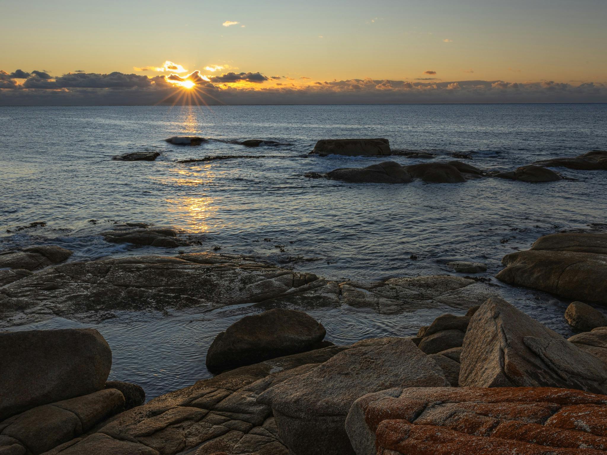 rocky coastline in foreground, sun on the horizon at sunrise