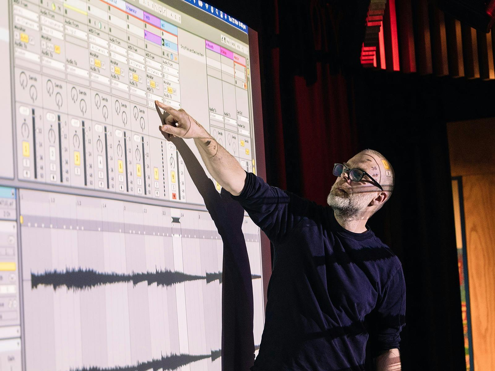 Man in glasses pointing at screen that is projecting an electronic sound desk.