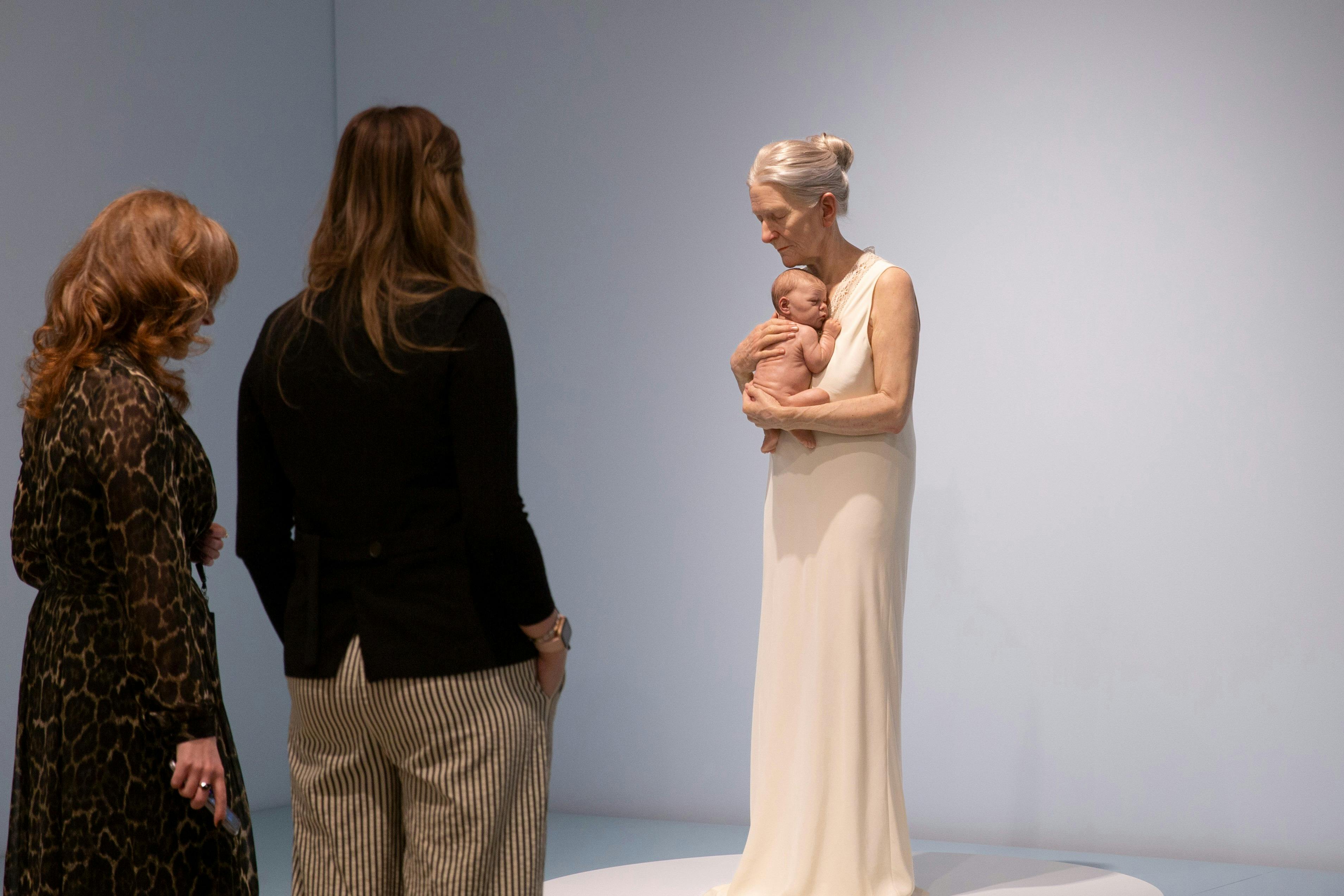 Two women view a hyperrealist sculpture of an elderly woman embracing a newborn baby