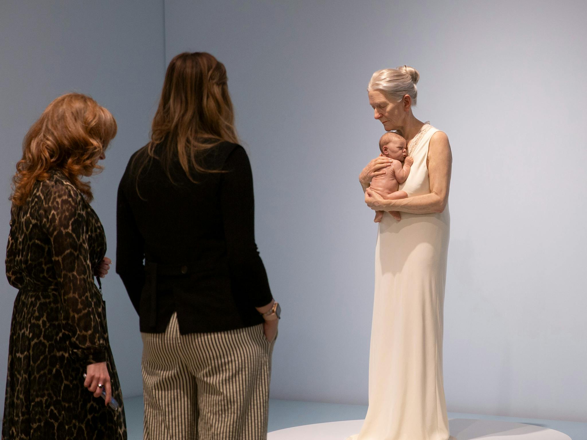 Two women view a hyperrealist sculpture of an elderly woman embracing a newborn baby