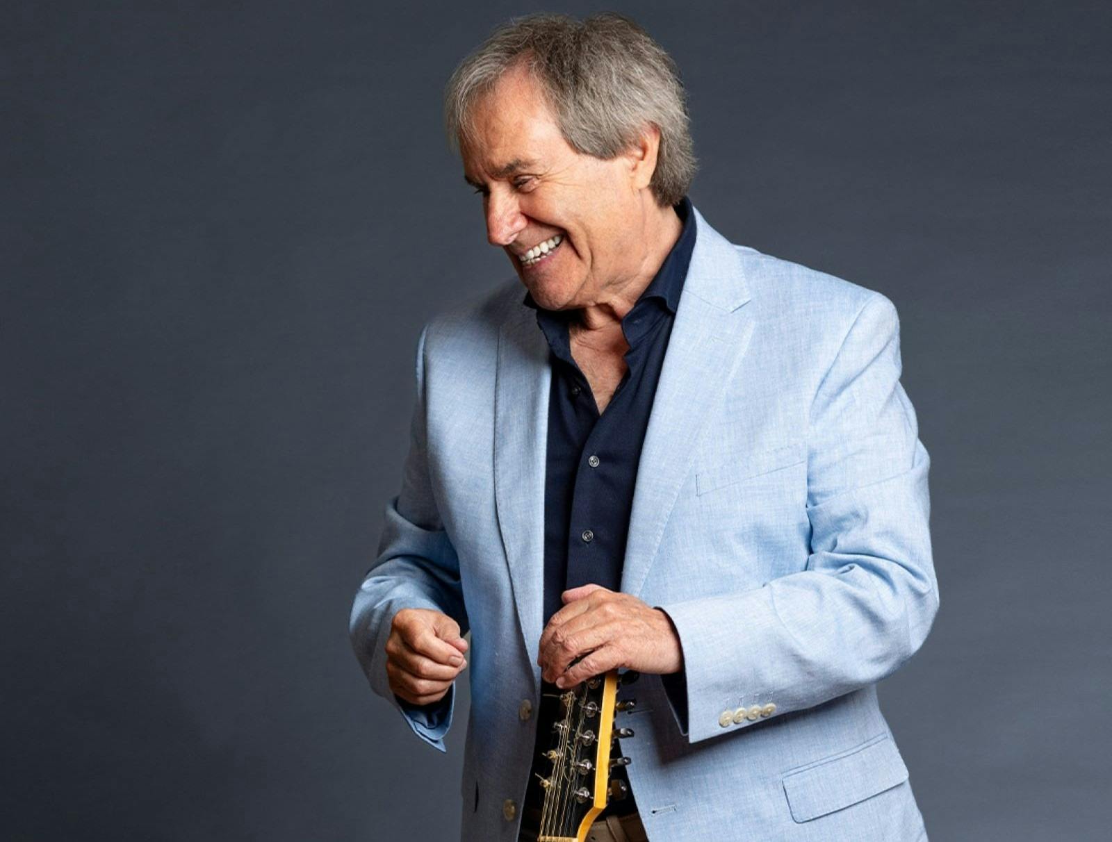 photo of Chris behind a grey background looking down and smiling holding a guitar