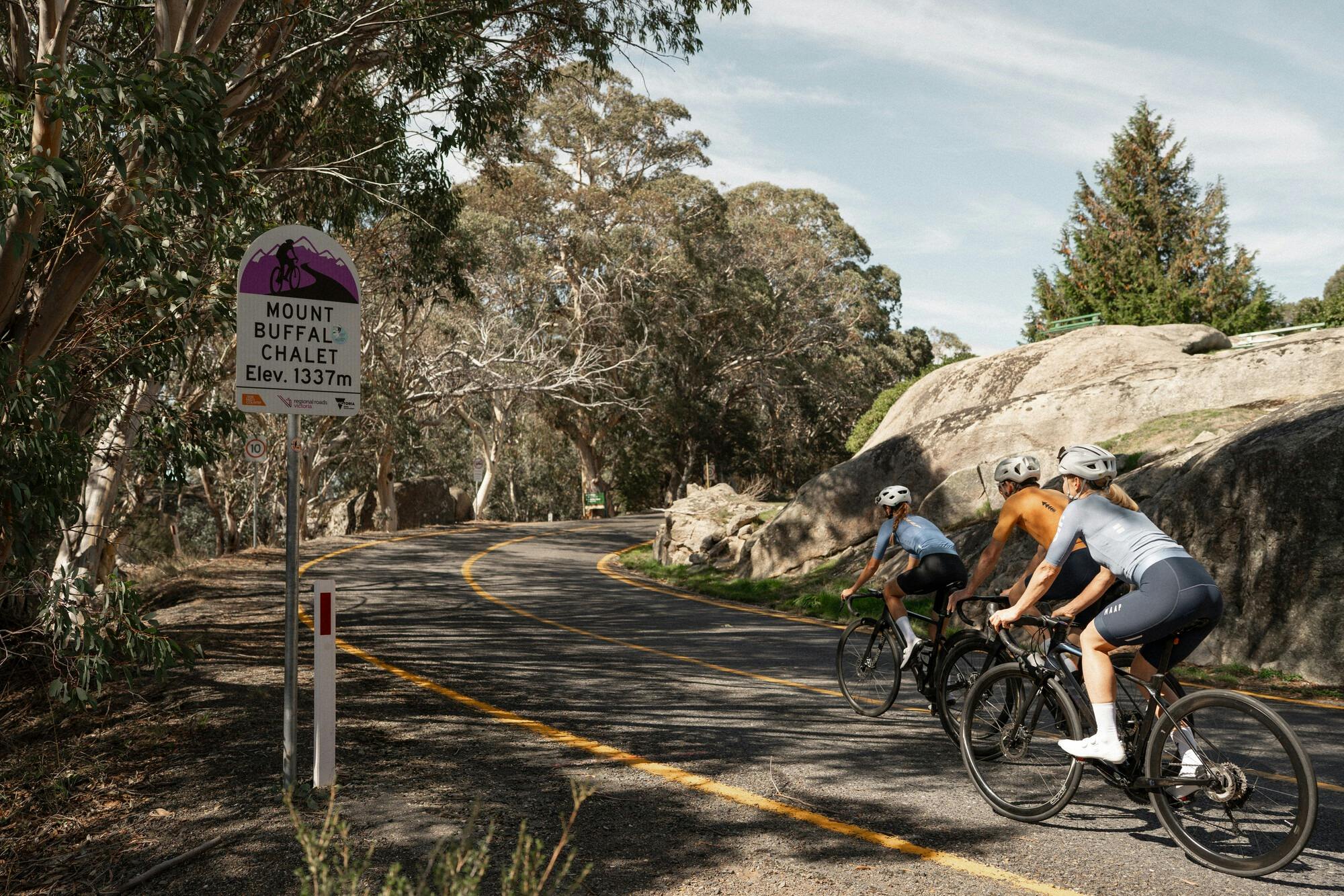 Three cyclists riding past the 7 Peaks Challenge sign on the Mount Buffalo 7 Peaks Challenge route