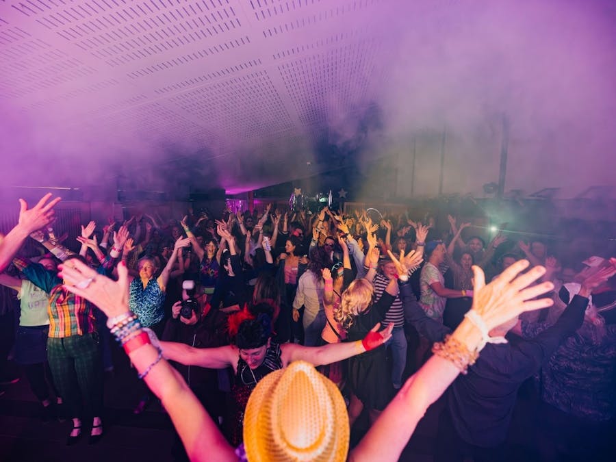 A performer throws their hands up in the air in a hall filled with dancers, purple lights and smoke