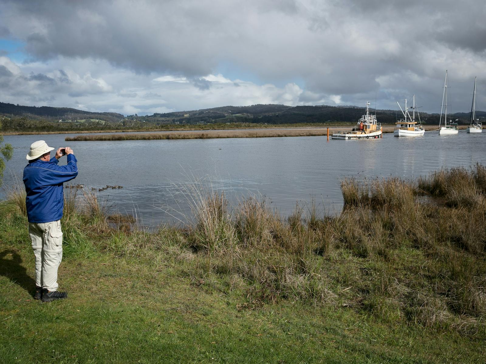 Guest standing at the edge of the Huon River pointing camera to boats on the water