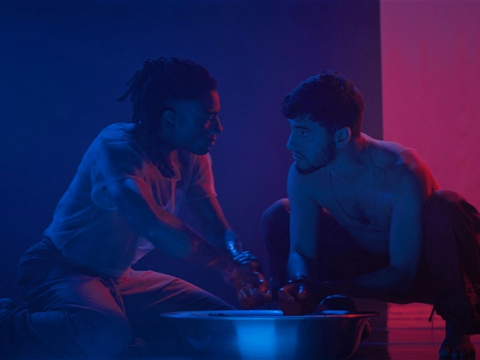 Two performers kneel facing each other with hands meeting in a basin of water lit in deep blue & red