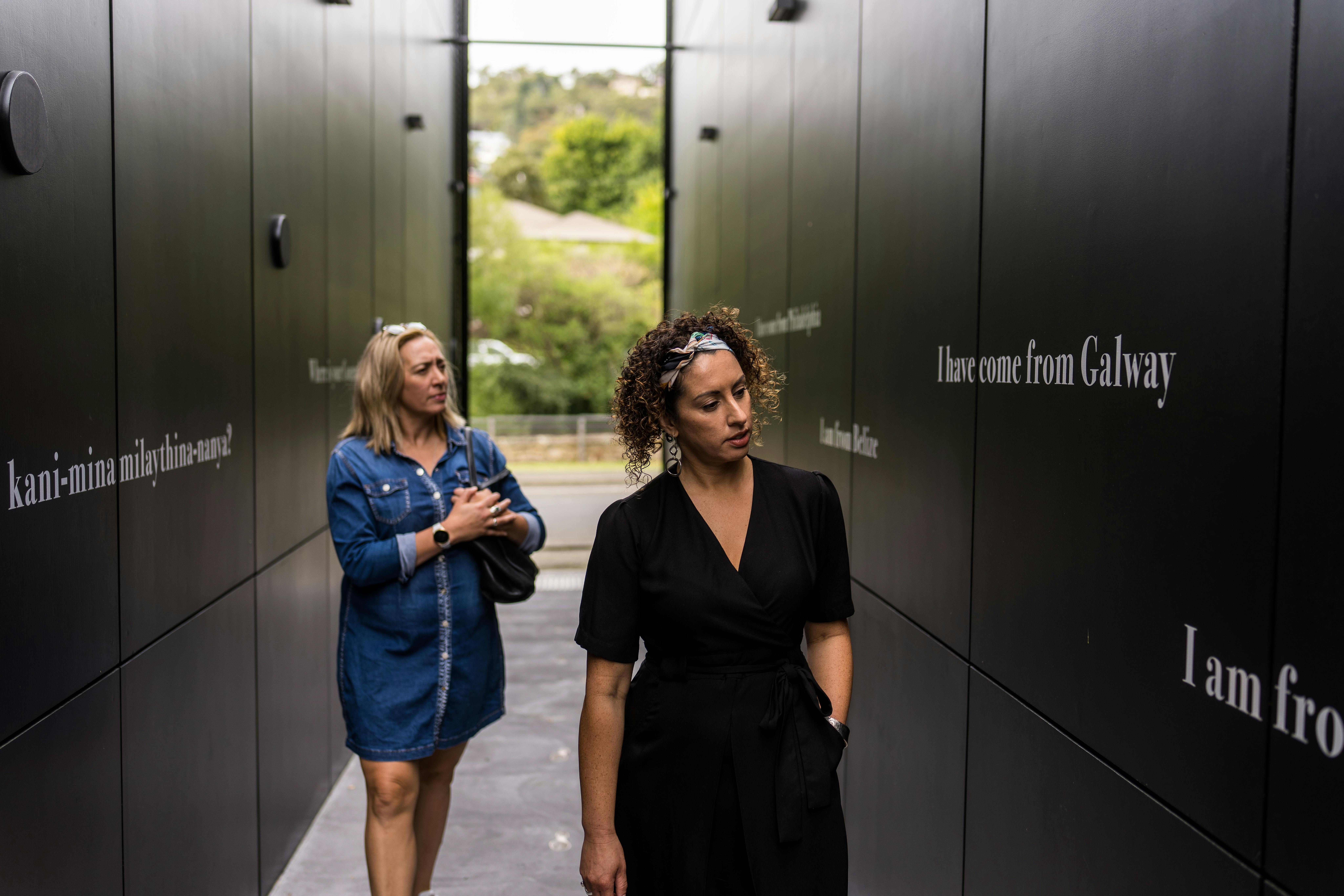 Two ladies walking through the entry walkway and reading the white texts  printed on the black walls