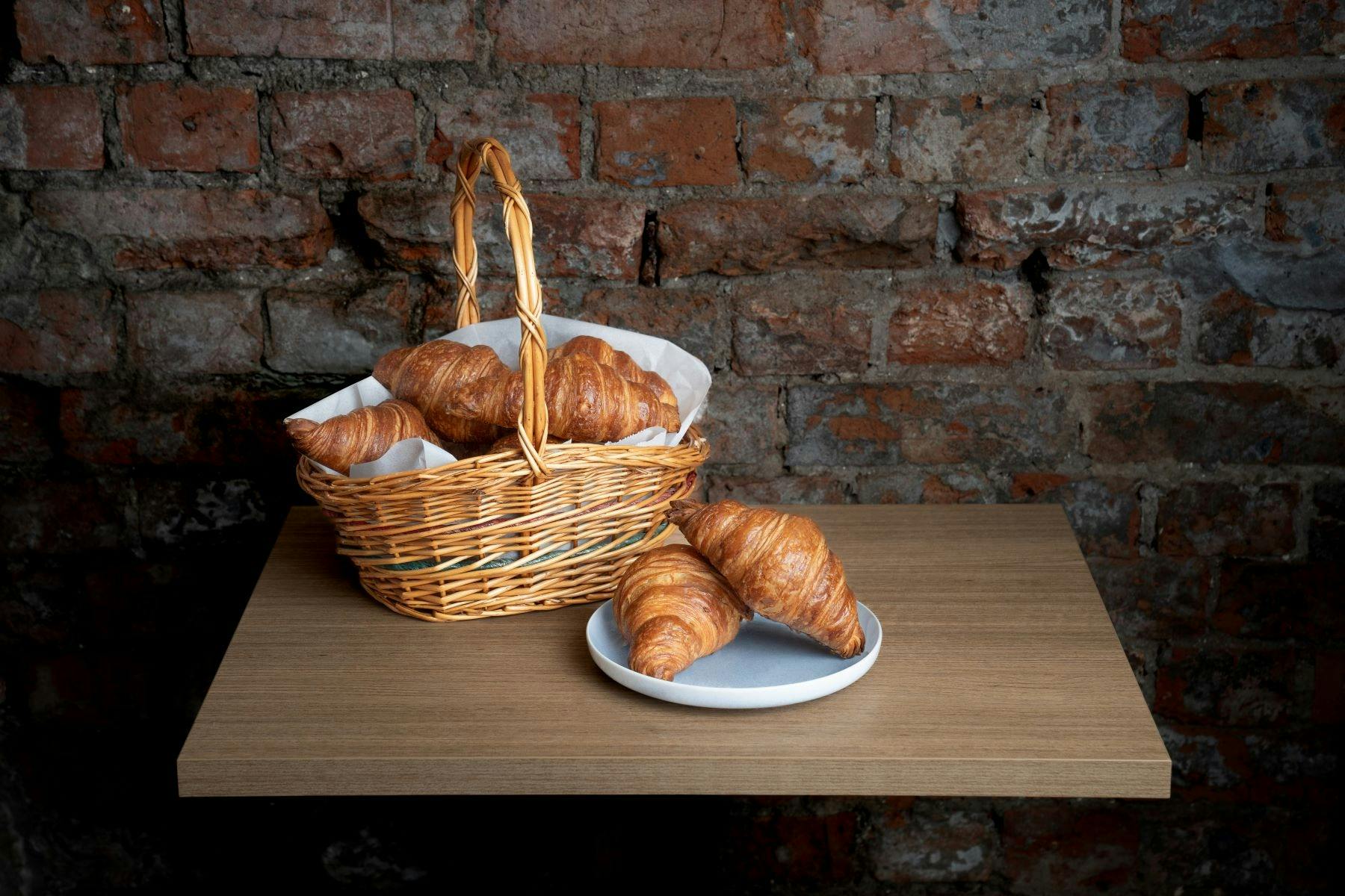 A basket of fresh croissants on a table against a rustic brick wall.
