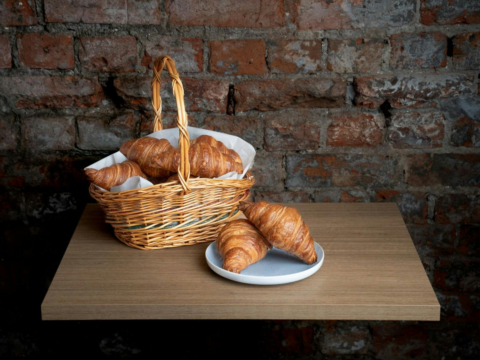 A basket of fresh croissants on a table against a rustic brick wall.