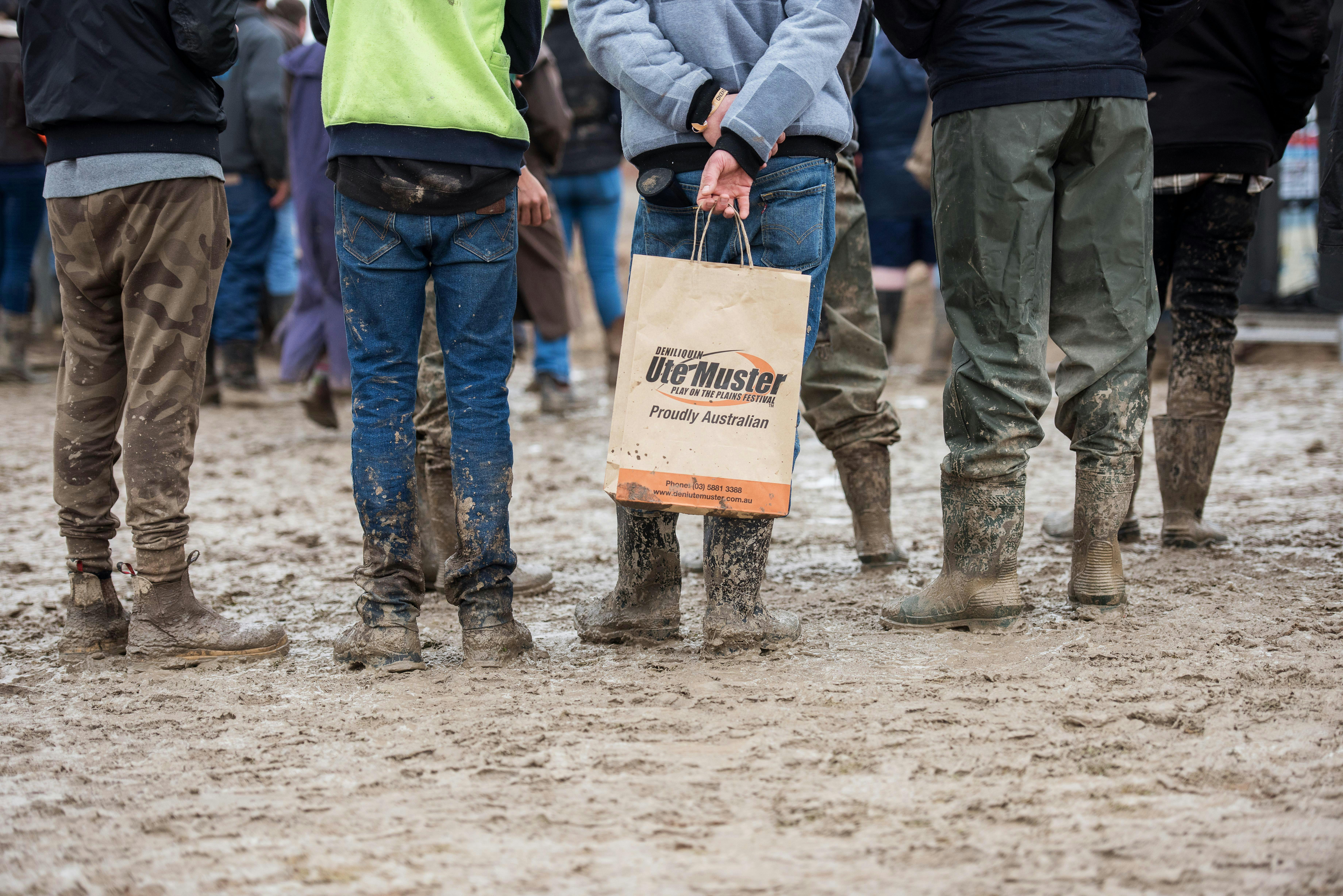 The backs of 5 people standing in the mud, one holds a Ute Muster bag behind their back.