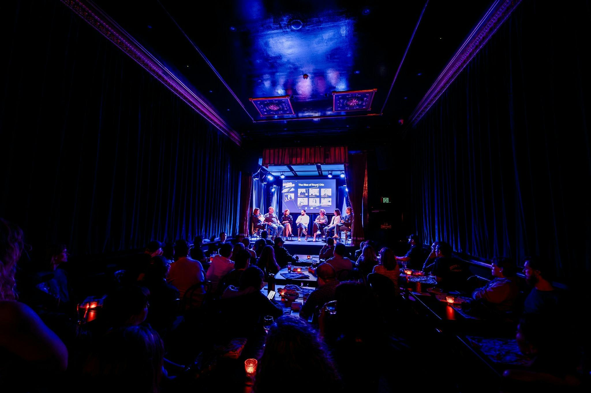 Members of a panel discussion sit on a stage while an audience watches from small tables
