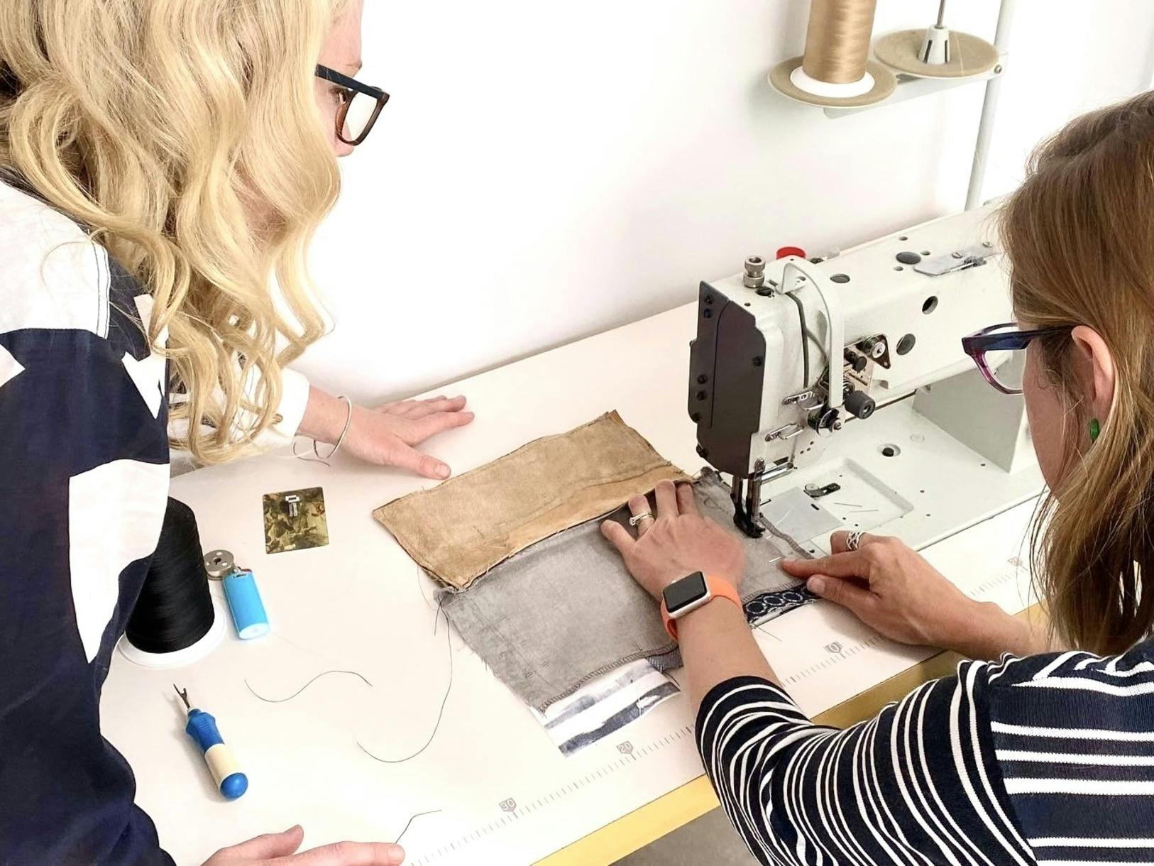 Two women working at a sewing machine during a leather workshop. One woman is guiding fabric under t