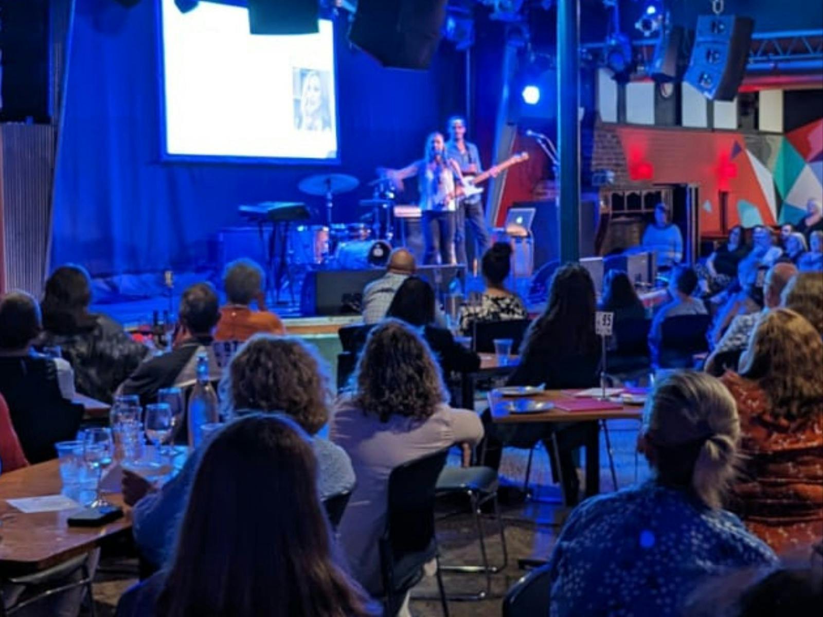 A crowd of people are sitting in a pub learning to sing a song in three part harmony