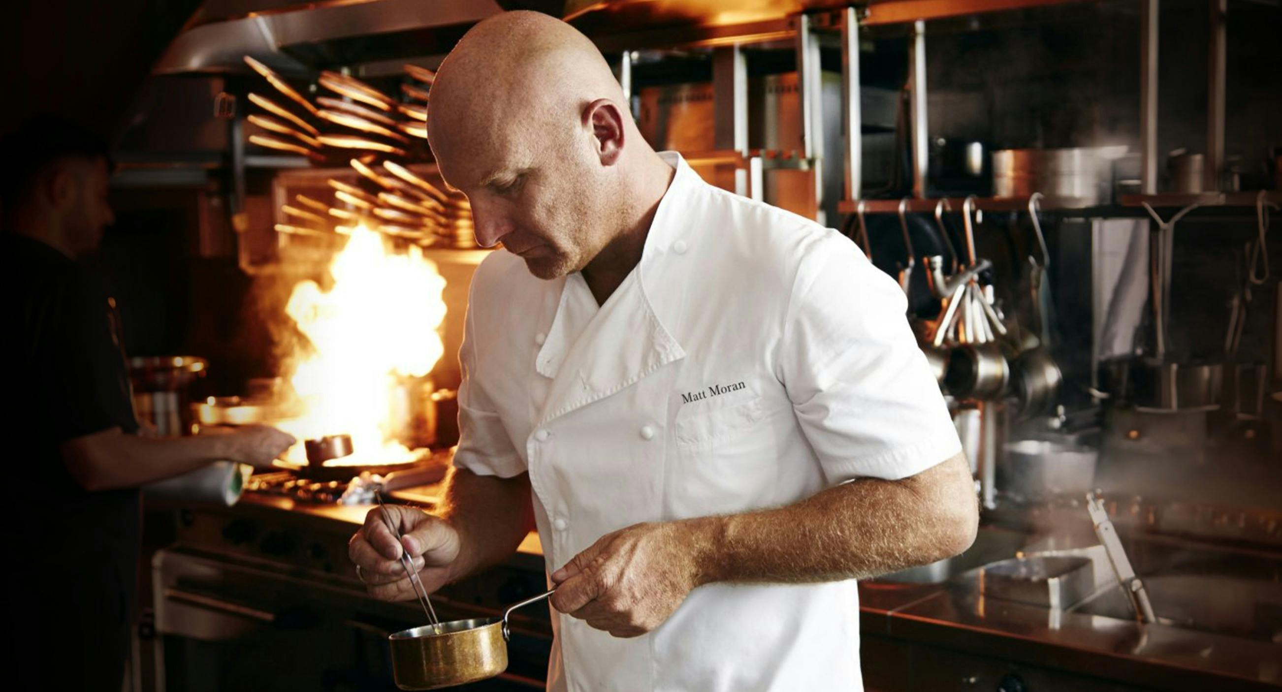 A man in a kitchen, stirring the contents of a small pot.