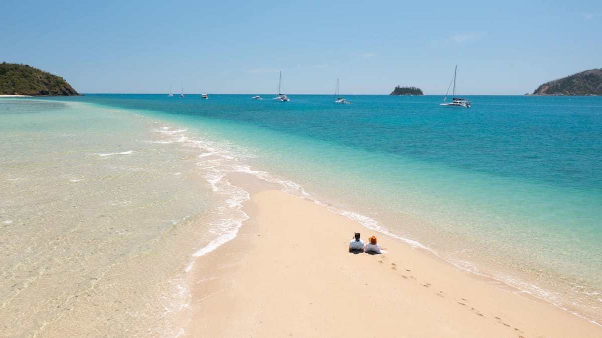 Two people laying on a sand stretch with several sailing boats moored in the backrgound