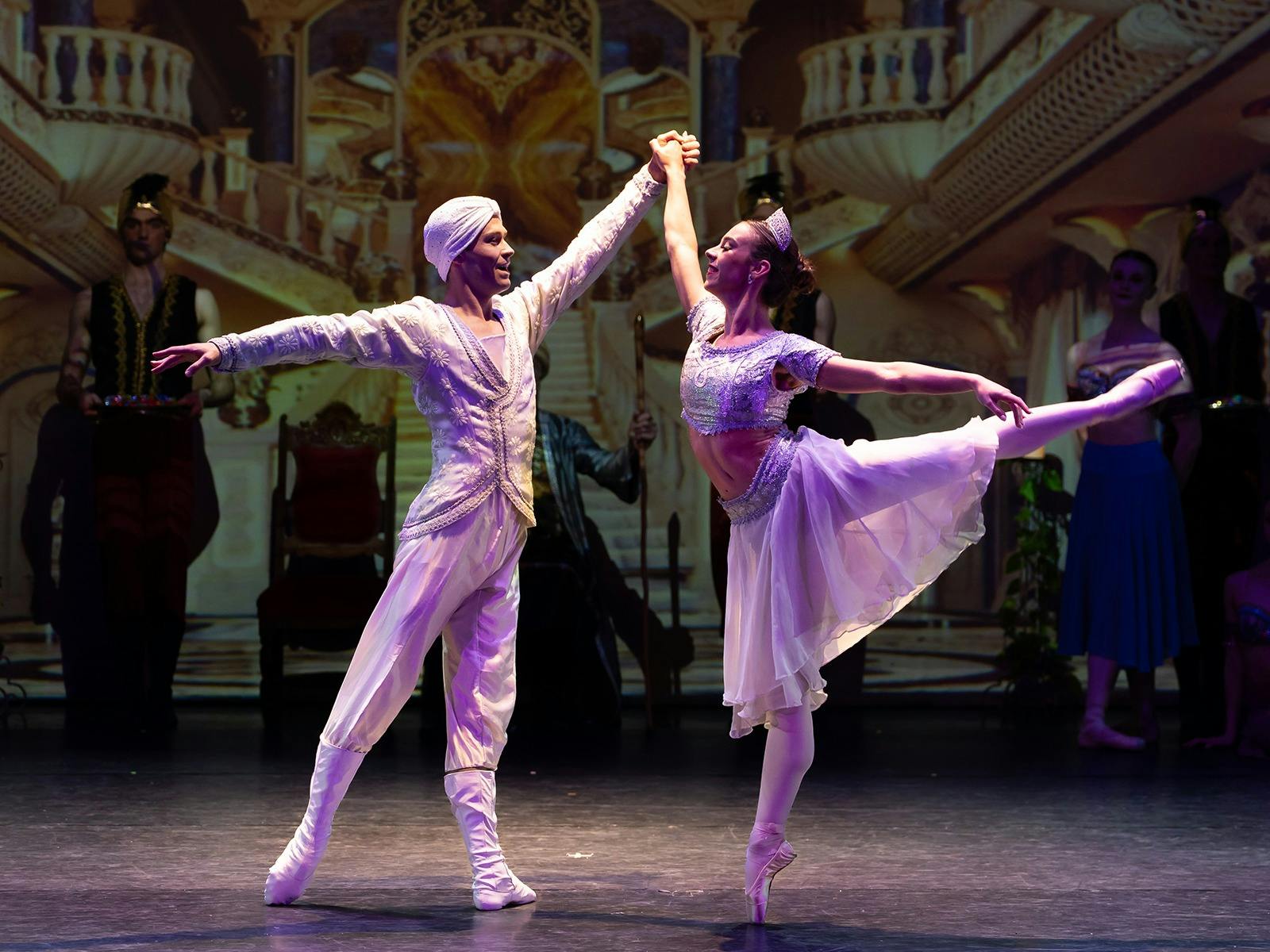 Two ballet dancers pose, one has their leg raised high in the air behind them