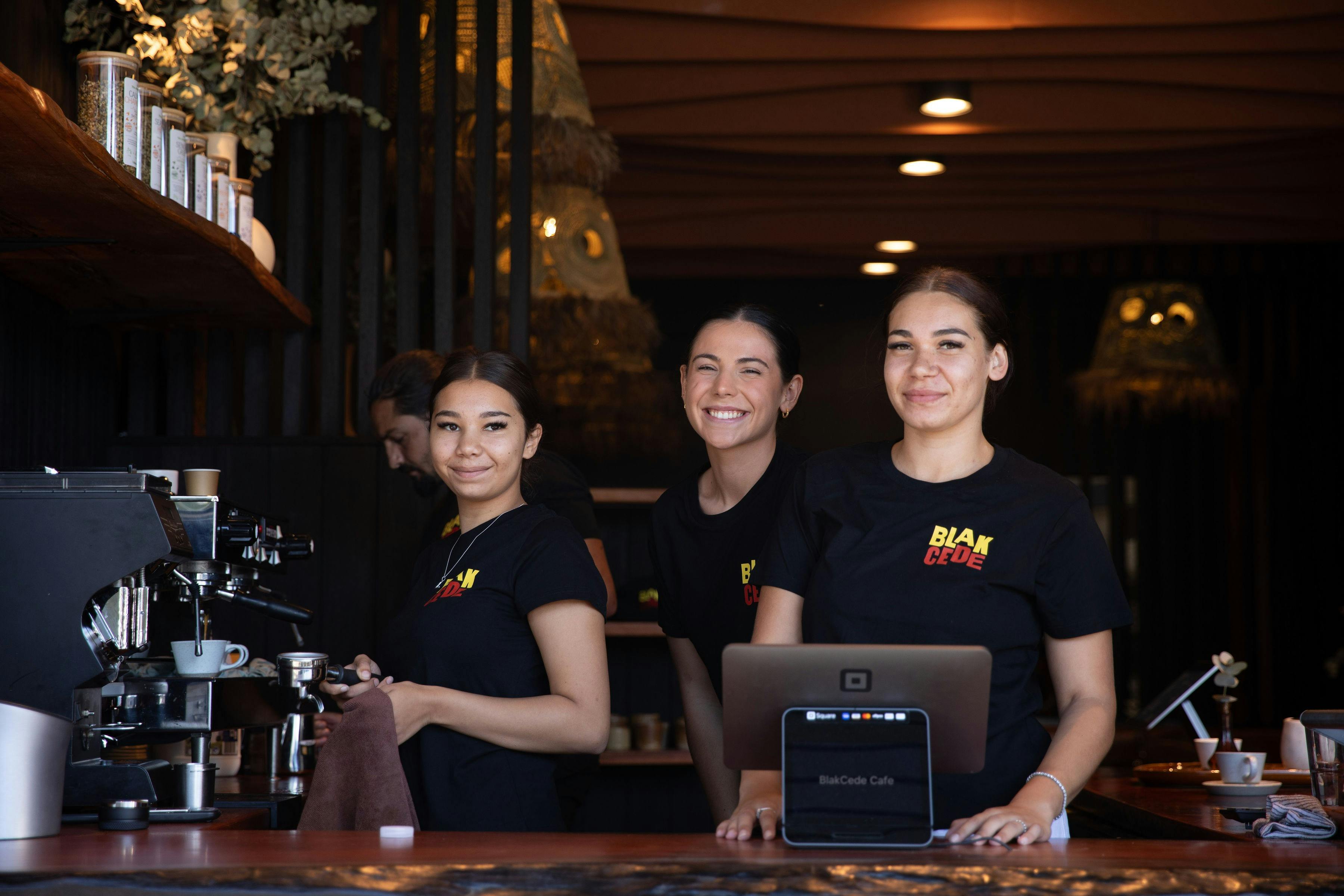 Three Blak Cede staff standing and smiling at coffee machine through window that opens up to street