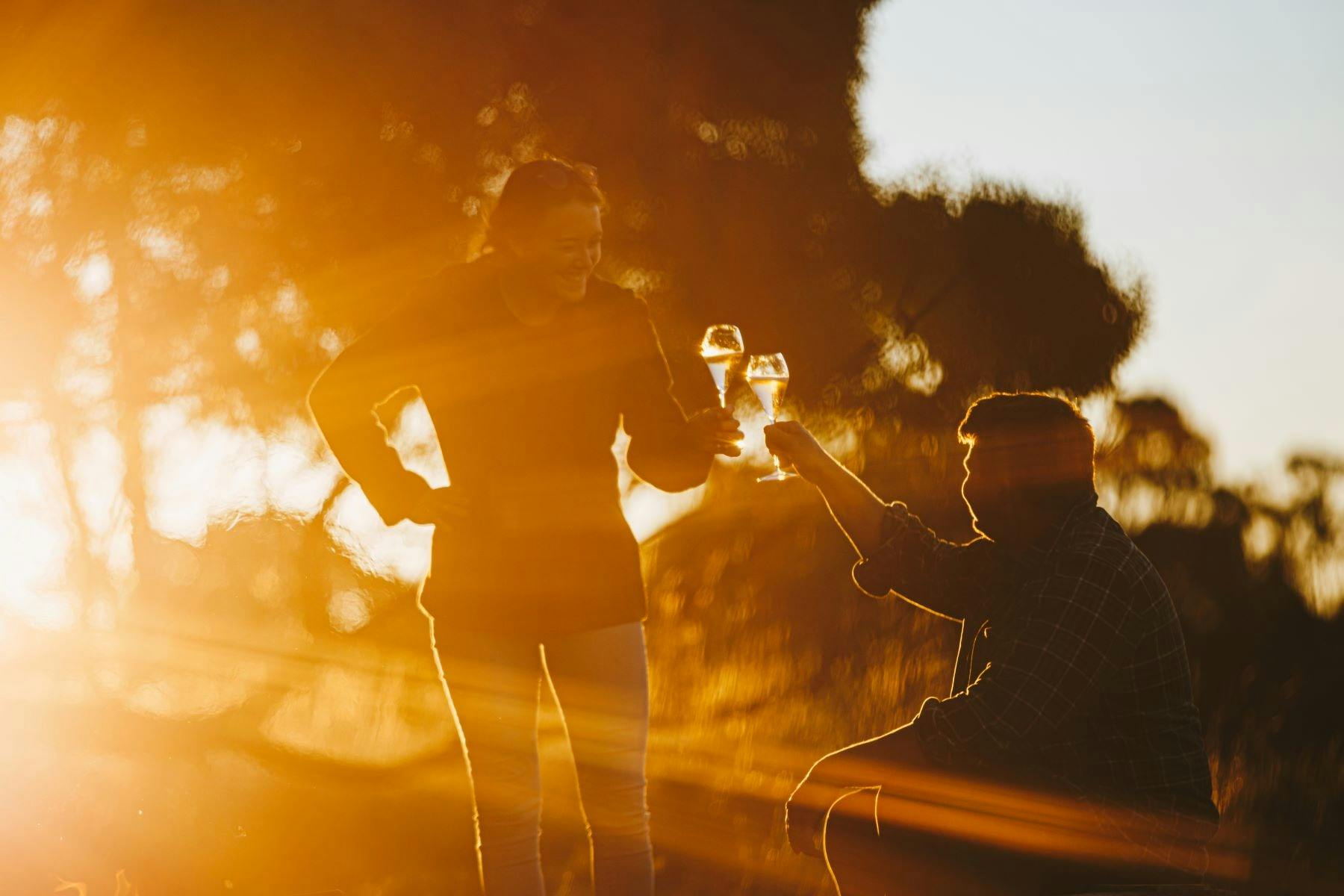 Man sitting and woman standing holding wine glasses with sun setting behind gum trees