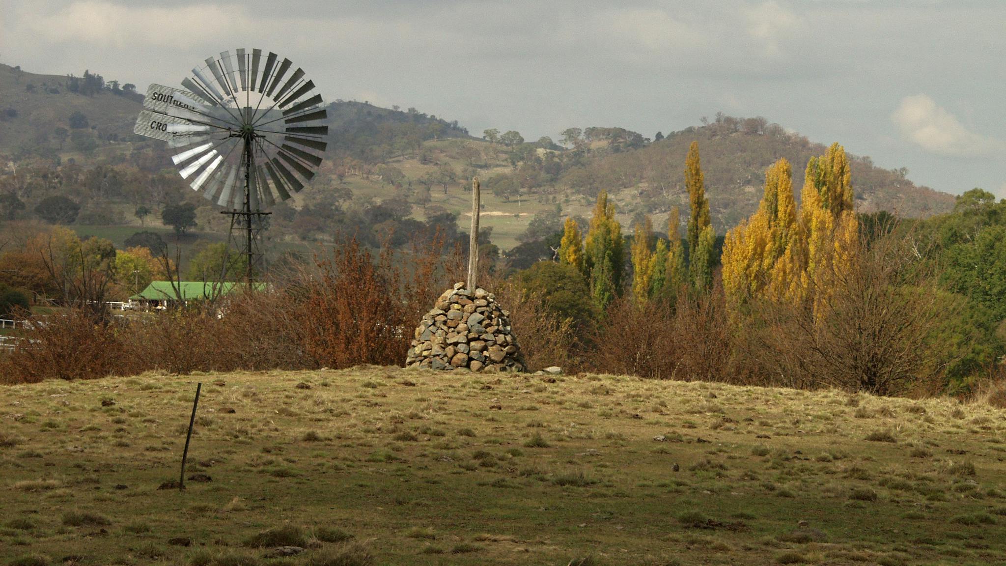 Grassy foreground with stone cairn, windmill and poplars and homestead