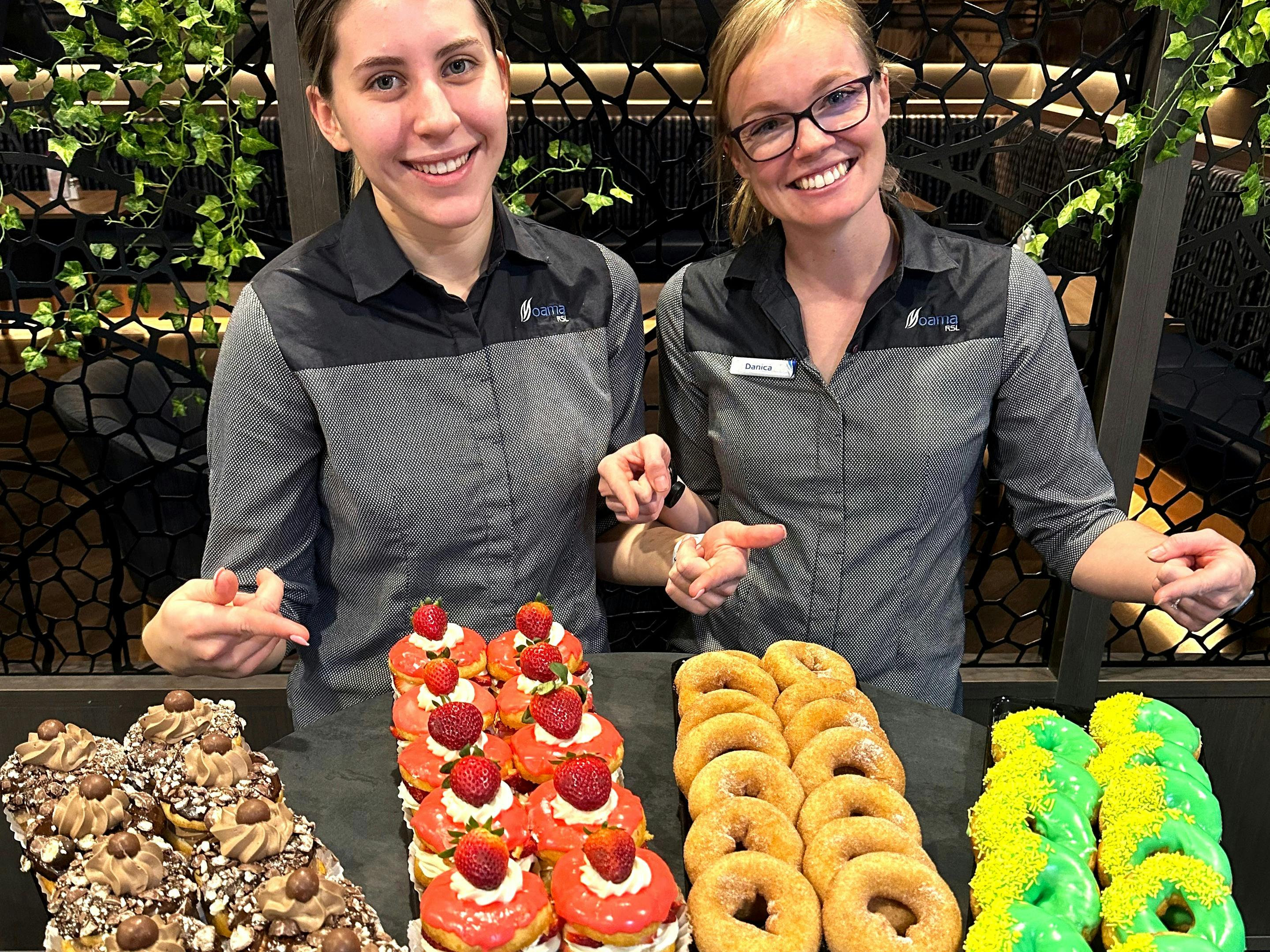 Two female staff members pointing to a variety of delicious donuts