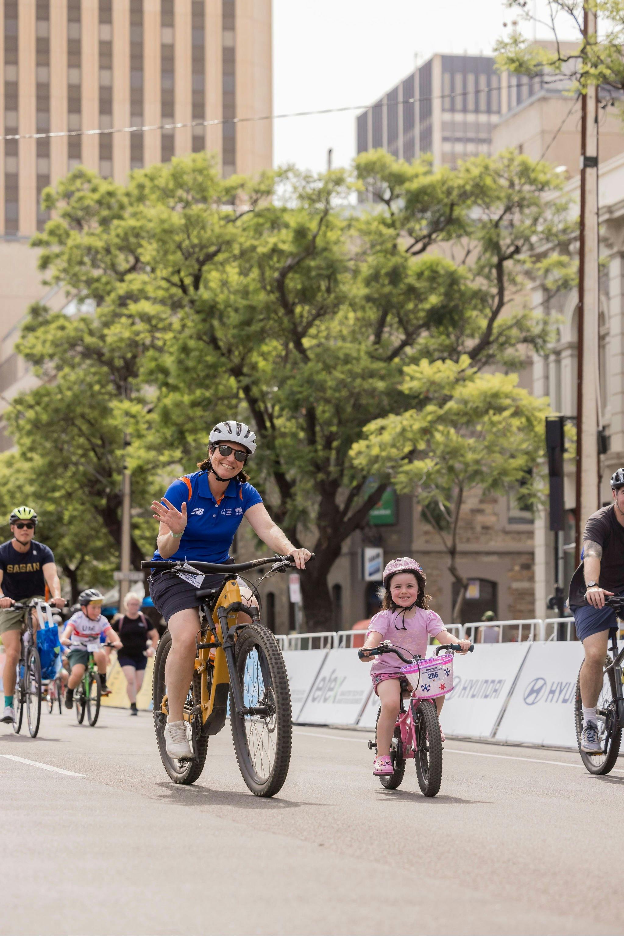 A women riding her bike smiling and waving alongside a young girl also riding her bike on a track.