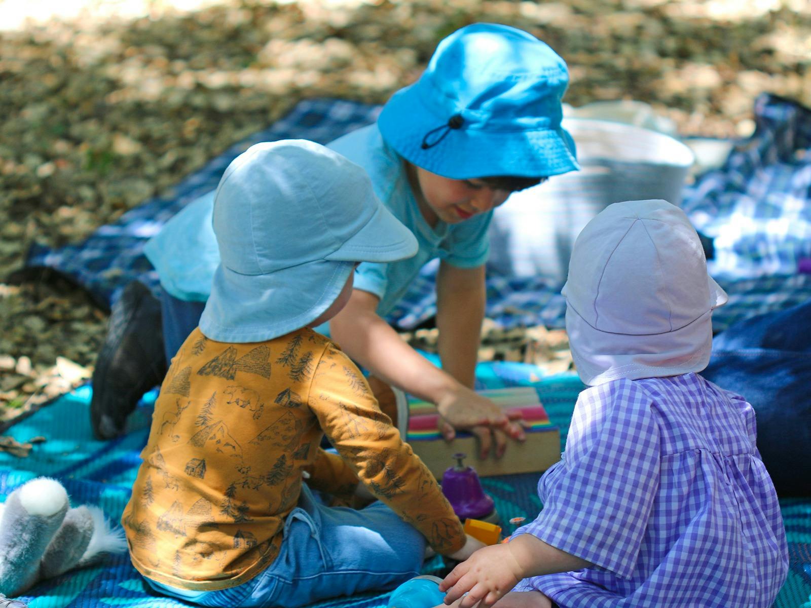 Three preschool children sit on picnic mats on the forest floor playing with wooden blocks