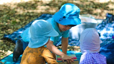 Three preschool children sit on picnic mats on the forest floor playing with wooden blocks