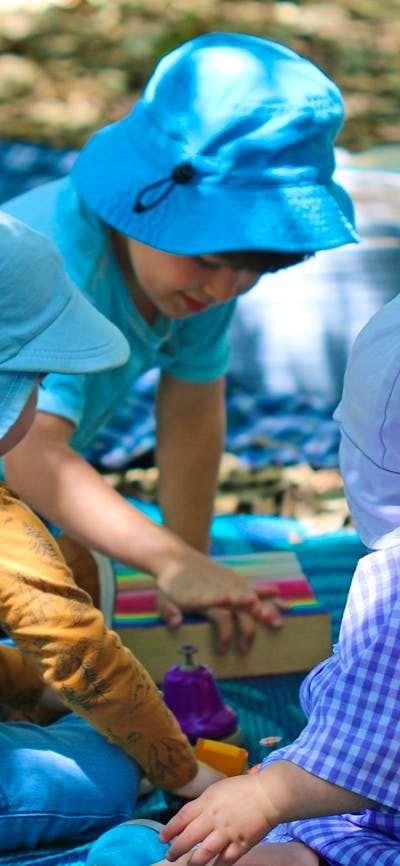 Three preschool children sit on picnic mats on the forest floor playing with wooden blocks