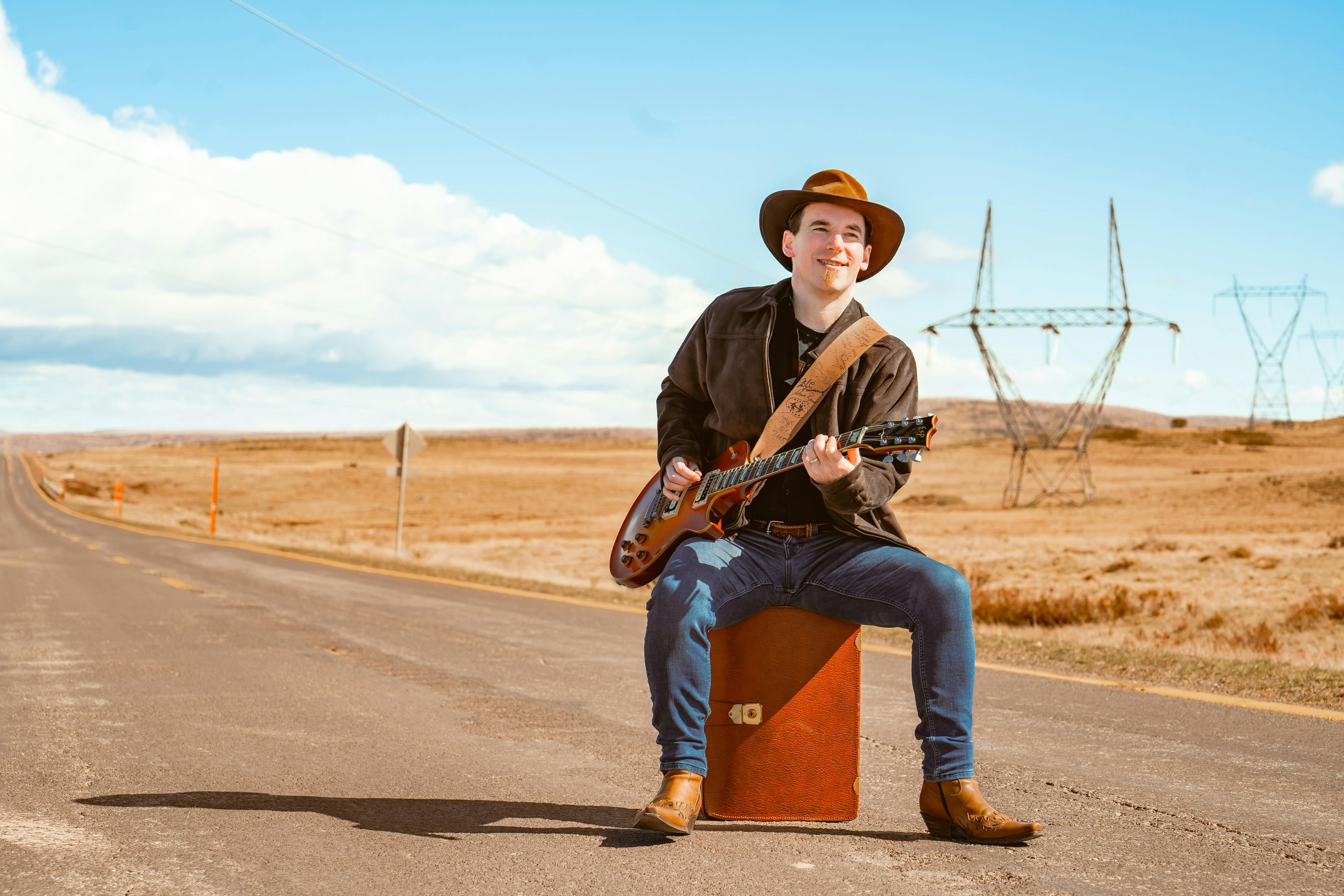 Man wearing an akubra sits on suitcase on highway while playing a guitar