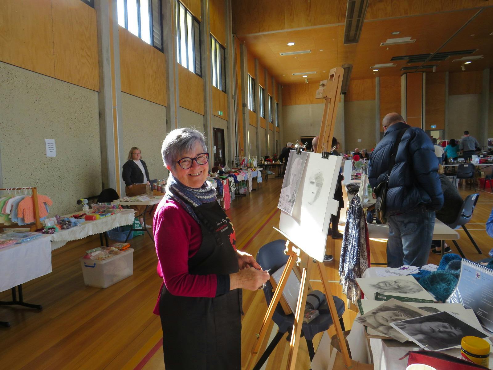 A lady in a dark pink jumper and black apron stands before a painting easel