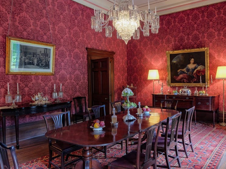 Deep red wallpaper covers the formal dining room. An ornate chandelier hangs from the ceiling.