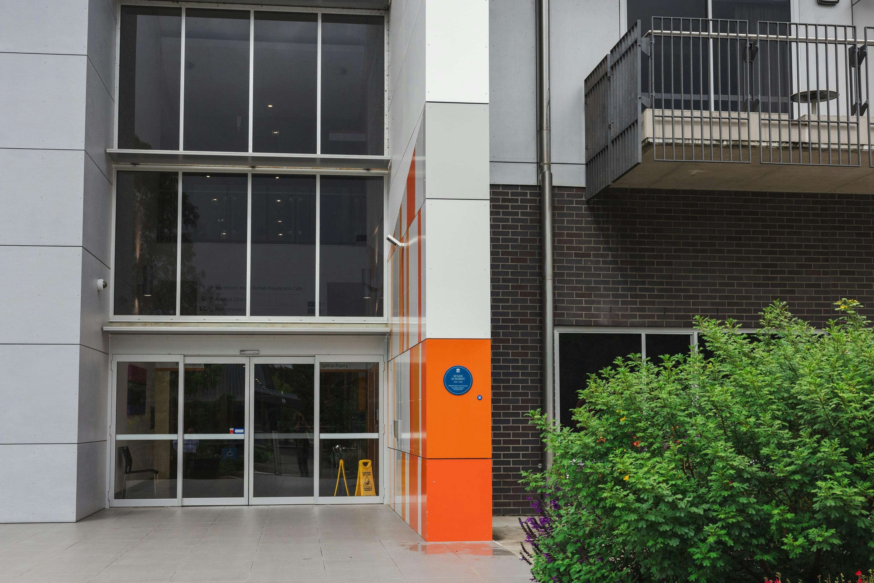 Blue plaque on orange section of wall next to entry doors of building with floor to ceiling windows